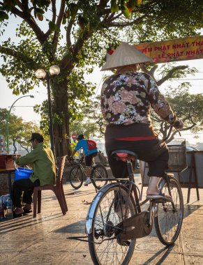 Geleneksel konik şapka takan bir kadın sabahın erken saatlerinde Hanoi, Vietnam 'daki Batı Gölü' nün yanında bisiklet sürüyor..