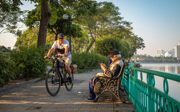 Старик сидит на скамейке и смотрит на свой мобильный телефон, как другой старик проезжает мимо ранним утром рядом с Вест-Лейк, Ханой, Вьетнам.