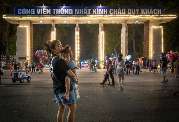 Молодая мать выносит свою маленькую дочь у входа в парк воссоединения в субботу вечером в Ханое, Вьетнам. Люди ходят, дети играют и женщины продают воздушные шары.