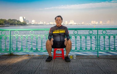 A middle aged Vietnamese man dressed in cycling gear rests on a Sunday morning next to the West Lake in Hanoi, Vietnam. clipart