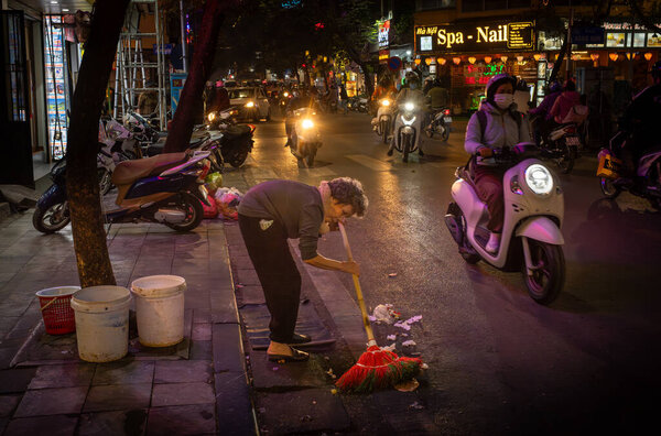 Пожилая вьетнамская женщина, страдающая кифозом, выметает мусор на обочине дороги в Ханг-Дье ночью в старом квартале, Ханой, Вьетнам.