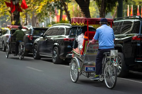 Bir bisikletli sürücü, Hanoi, Vietnam 'ın merkezindeki Hoan Kiem Gölü yakınlarında park halindeki arabaların yanından geçen bir turisti taşıyor.