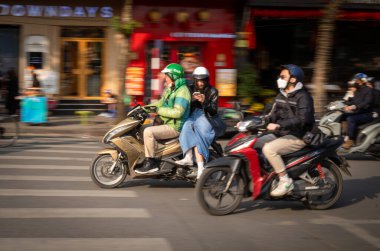 A young Vietnamese woman looks at her mobile smartphone as she rides side saddle on the back of a Grab motorcycle taxi next to Hoan Kiem Lake in central Hanoi, Vietnam clipart