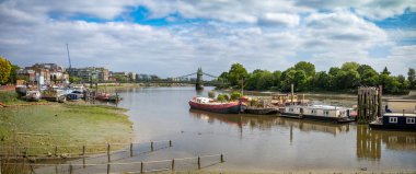 Viktorya dönemi Hammersmith Köprüsü yakınlarındaki Thames nehrine demirlemiş dönüştürülmüş mavnalar ve yüzen evlerden oluşan bir panorama..