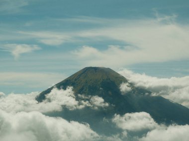 Bir dağ bulutlarla kaplıdır ve uzaktan görülebilir. Gökyüzü çoğunlukla mavi ve bazı beyaz bulutlar etrafa saçılmış. Dağ dik dururken manzara sakin ve huzurlu.