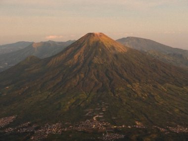 Tepesinde yeşil bir orman ve altında bir şehir olan bir dağ. Dağ ağaçlarla kaplı ve şehir uzakta.