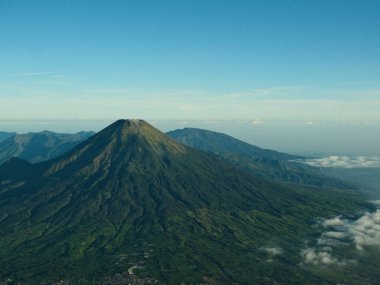Ortasında büyük yeşil bir dağ olan bir dağ sırası. Gökyüzü açık ve mavi. Dağ ağaçlarla kaplı, yemyeşil ve yemyeşil.