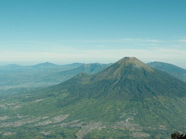 Yeşil bir ormanı ve arka planda bir şehri olan bir dağ sırası. Gökyüzü açık ve mavi