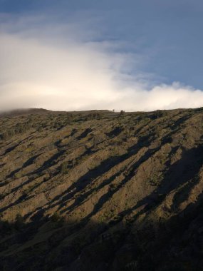 Bulutlu bir gökyüzü ve birkaç ağacı olan bir dağ. Bulutlar beyaz ve gökyüzü mavi
