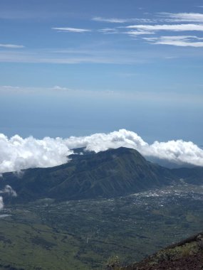 Arkasında bulutlu bir gökyüzü olan bir dağ. Bulutlar beyaz ve kabarık. Dağ yeşil bir manzarayla çevrilidir.