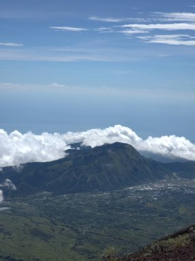 Arkasında bulutlu bir gökyüzü olan bir dağ. Bulutlar beyaz ve kabarık. Dağ yeşil bir manzarayla çevrilidir.