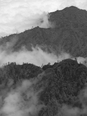 Görüntü, gökyüzünü kaplayan bulutlu bir dağ silsilesinin siyah beyaz fotoğrafı. Bulutlar sükunet ve sükunet hissi yaratırken manzara sakin ve huzurlu.