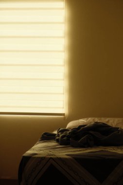 Battaniyeli bir yatak ve arka planda bir pencere. Oda loş ve yatak dağınık.