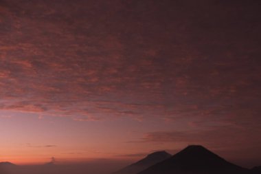 Pembe ve turuncu gökyüzüne karşı bir dağ silueti var. Dağlar arka planda ve gökyüzü bulutlarla dolu.