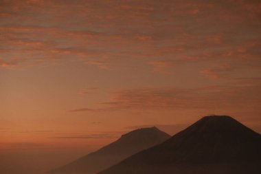 Bir dağ silueti turuncu ve pembe gökyüzünün arka planına karşı. Dağlar sisli bir sis ile kaplıdır. Sahneye rüya gibi, ruhani bir hava katar.