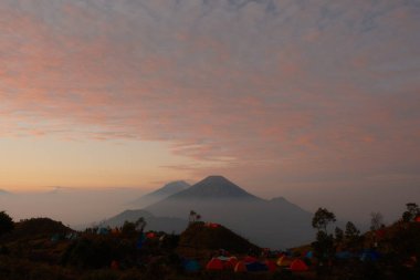 Arkasında güzel bir günbatımı olan bir dağ sırası. Gökyüzü bulutlarla dolu ve güneş batıyor. Dağlar ağaçlarla çevrili ve her tarafta çadırlar var.