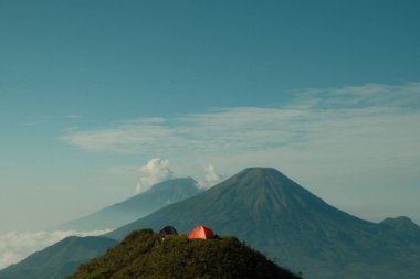 Dağın tepesinde kırmızı bir çadır var. Gökyüzü mavi ve bulutlar var. Dağlar arka planda.