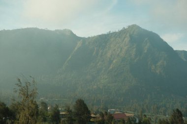 Uzakta bir kasabası olan bir dağ sırası. Gökyüzü bulutlu ve dağlar ağaçlarla kaplı.