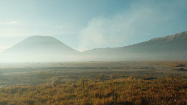 Ortasında büyük bir volkan olan bir dağ sırası. Gökyüzü açık ve mavi. Dağlar ağaçlarla kaplı ve volkan dumanla kaplı.