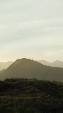 Ortasında küçük bir tepe olan bir dağ sırası. Gökyüzü bulutlu ve güneş batıyor.