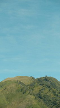 Arka planda açık mavi gökyüzü olan bir dağ. Gökyüzü çok parlak ve dağ çok yeşil.