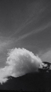 Tepesinde bulut olan bir dağ. Bulut beyaz ve kabarık. Gökyüzü karanlık ve bulutlu.