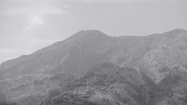 Arka planda bulutlu bir gökyüzü olan bir dağ sırası. Dağlar ağaçlarla çevrili, gök de bulutlarla çevrili.