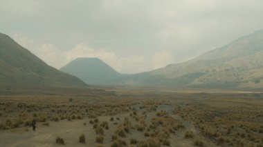 Arka planda bir dağ olan bir insan çölde yürüyor. Gökyüzü bulutlu ve manzara kuru ve çorak.