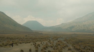 Arka planda bir dağ olan bir insan çölde yürüyor. Gökyüzü bulutlu ve manzara kuru ve çorak.