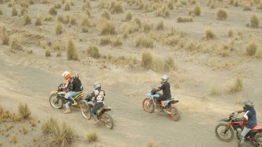 Toprak bir yolda dört kişi motosiklet sürüyor. Biniciler kask ve sırt çantası takıyor. Sahne maceralı ve heyecan verici.