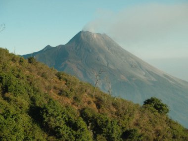 Yeşil yamacı ve bulutlu gökyüzü olan bir dağ. Dağ ağaçlarla kaplı ve uzakta bir ateş var.