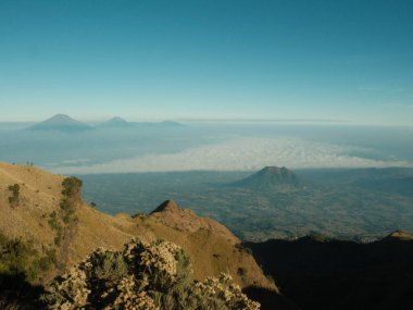 Arka planda bulutlu bir gökyüzü olan bir dağ sırası. Dağlar ağaçlarla kaplı ve gökyüzü mavi