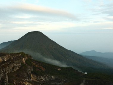 Üstünde sis bulutu olan bir dağ. Dağ yeşil ve gökyüzü mavi.