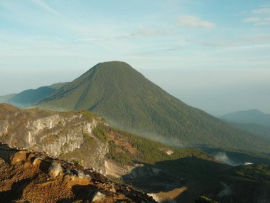 Tepesinde yeşil orman olan bir dağ. Gökyüzü mavi ve güneş parlıyor. Dağ çok yüksek ve ağaçlar yemyeşil.