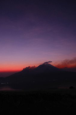 Bir dağ güzel bir günbatımının arka planındadır. Gökyüzü koyu mor renktedir ve dağ dumanla kaplıdır.
