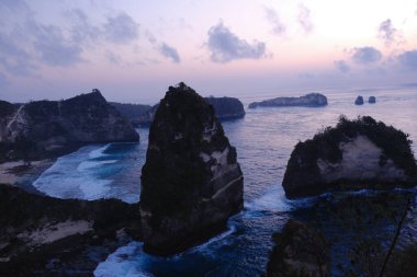 Arka planda güzel bir günbatımı olan kayalık bir kıyı şeridi. Su sakindir ve gökyüzü turuncu ve mor renklerin karışımıdır.