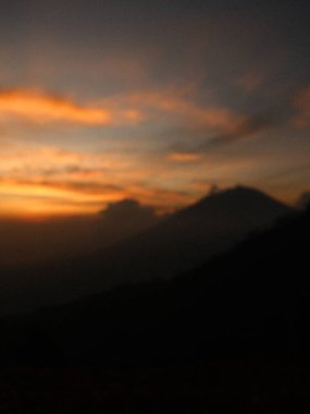 Arka planda gün batımı olan bir dağ silsilesinin bulanık fotoğrafı. Gökyüzü turuncu ve dağlar gökyüzüne karşı siluetlenmiş.