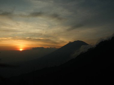 Arka planda güneş batan karanlık bir gökyüzüne karşı bir dağ silueti var. Gökyüzü bulutlu ve güneş batıyor, kasvetli bir atmosfer yaratıyor.