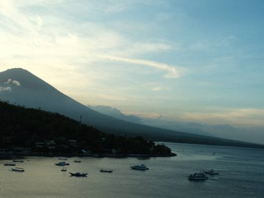 Arka planda dağ olan güzel bir göl manzarası. Gökyüzü bulutlu ve güneş batıyor. Suda çok sayıda tekne var, bazıları küçük, bazıları da büyük.