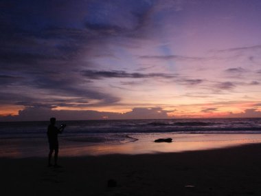 Sahilde elinde kamerayla duran bir adam güzel gün batımının fotoğrafını çekiyor. Gökyüzü bulutlarla dolu ve güneş batıyor. Sıcak ve huzurlu bir atmosfer yaratıyor.