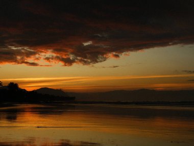 Sakin bir su üzerinde güzel bir gün batımı. Gökyüzü bulutlarla dolu ve güneş batıyor, sahne üzerine sıcak bir parıltı saçıyor. Su durgun ve huzurlu, sakin bir atmosfer yaratıyor.