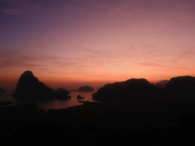 Bir dağ sırasının üzerinde suda bir tekneyle güzel bir gün batımı. Gökyüzü turuncu ve mor renklerin karışımıdır.