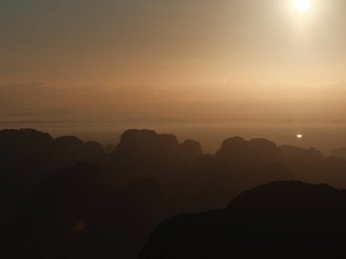 Arka planda güneş olan bir dağ silsilesinin bulanık fotoğrafı. Gökyüzü turuncu ve mavinin bir karışımıdır. Sıcak ve huzurlu bir atmosfer yaratır.