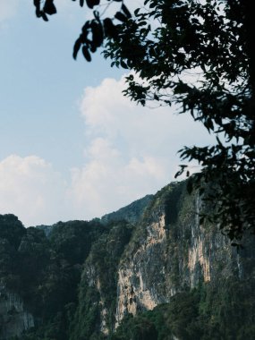 Arka planda bulutlu bir gökyüzü olan bir dağ sırası. Ağaçlar yemyeşil, gökyüzü mavidir.