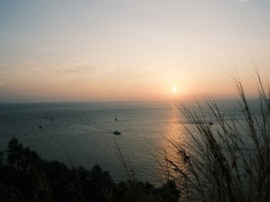 Güneş okyanusun üzerinde batıyor, suyun üzerine sıcak bir parıltı saçıyor. Gökyüzü mavi ve turuncunun bir karışımıdır. Huzurlu ve huzurlu bir atmosfer yaratır. Sudaki tekneler küçük ve dağınık.