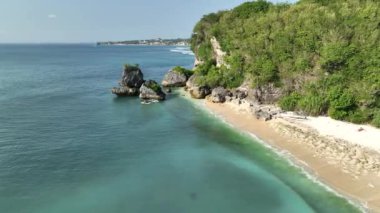 Küçük bir adası olan güzel bir tropikal sahil manzarası. Beyaz kum turkuazı okyanus ve kayalar. Kumsaldaki güneş pansiyonları. Thomas Beach Endonezya Bali. 4K dji Maviç 3 video. 