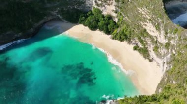 İnsanlar güneşleniyor ve güzel tropikal ada sahillerinde yürüyorlar. Arkasında kayalıklar olan beyaz kum ve turkuaz okyanus. Kelingking Plajı Nusa Penida Bali Endonezya. Hava aracı görüntüleri 4K