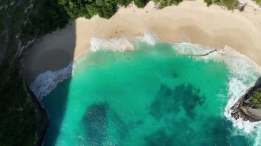 İnsanlar güneşleniyor ve güzel tropikal ada sahillerinde yürüyorlar. Arkasında kayalıklar olan beyaz kum ve turkuaz okyanus. Kelingking Plajı Nusa Penida Bali Endonezya. Hava aracı görüntüleri 4K