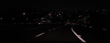 Gece, üzerinde beyaz şerit işaretleri olan karanlık dolambaçlı bir yol. Sokak lambaları yolu aydınlatıyor. Şehir ışıkları uzak tepelerde parıldıyor. Gece sürüşü, şehir manzarası veya ulaşım konuları için mükemmel..