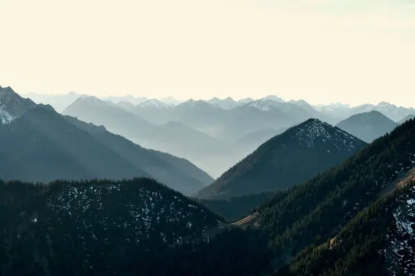 stock image Mesmerizing view of a misty mountain range at dawn, with layers of peaks fading into the distance. Soft morning light casting gentle shadows giving depth for the majestic landscape. 